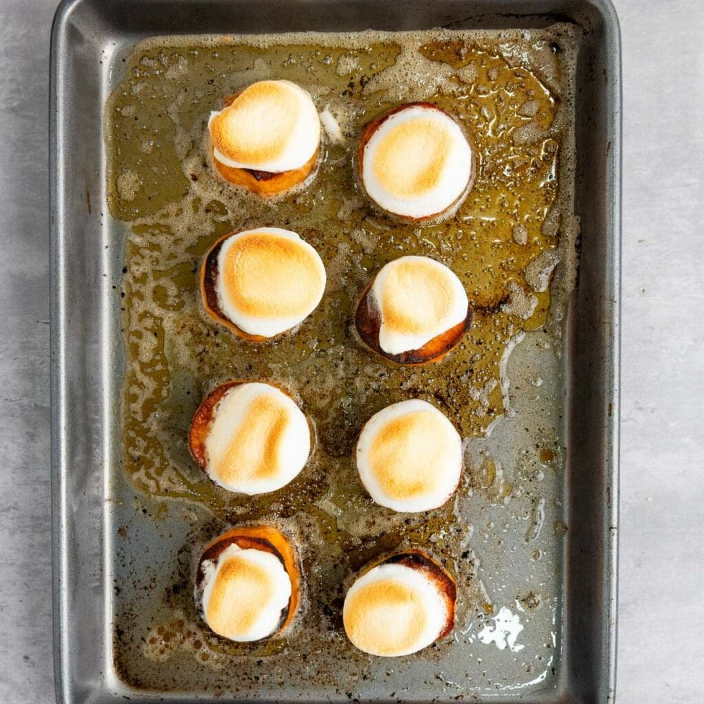 A baking tray with eight sweet potato rounds topped with toasted marshmallows, resting in melted butter on a gray surface.