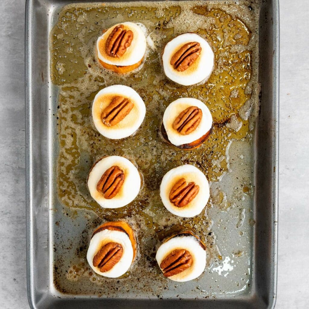 A baking tray with nine sweet potato rounds topped with melted marshmallows and pecan halves.