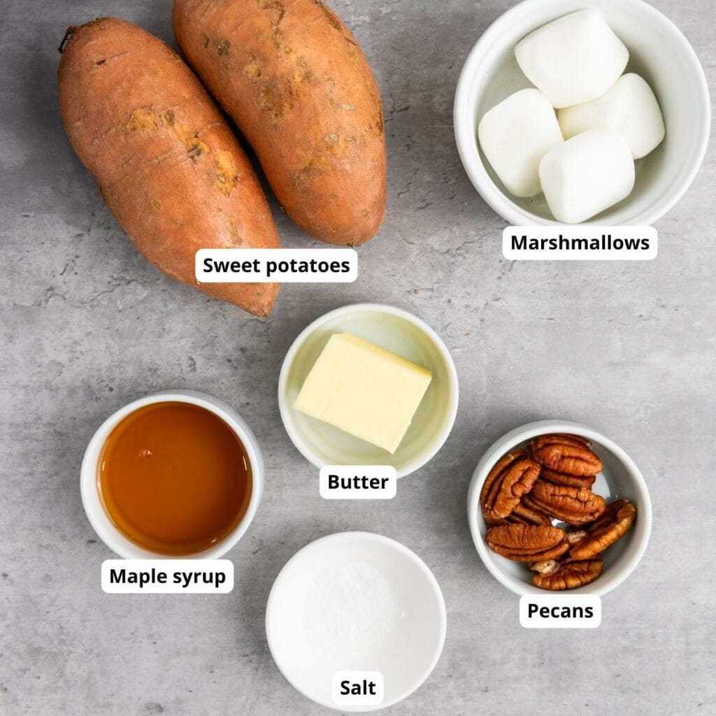 Two whole sweet potatoes, marshmallows, butter, maple syrup, salt, and pecans are arranged in bowls on a gray surface.