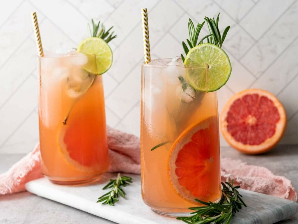 Two tall glasses of grapefruit cocktail with ice, garnished with lime slices, rosemary sprigs, and paper straws.