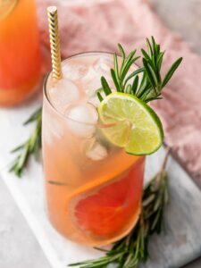 A tall glass of iced pink grapefruit drink garnished with a lime slice, rosemary sprig, and a striped straw, placed on a light surface.