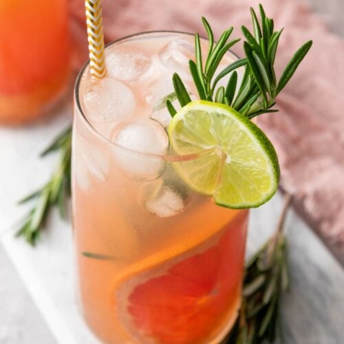 A tall glass of iced pink grapefruit drink garnished with a lime slice, rosemary sprig, and a striped straw, placed on a light surface.