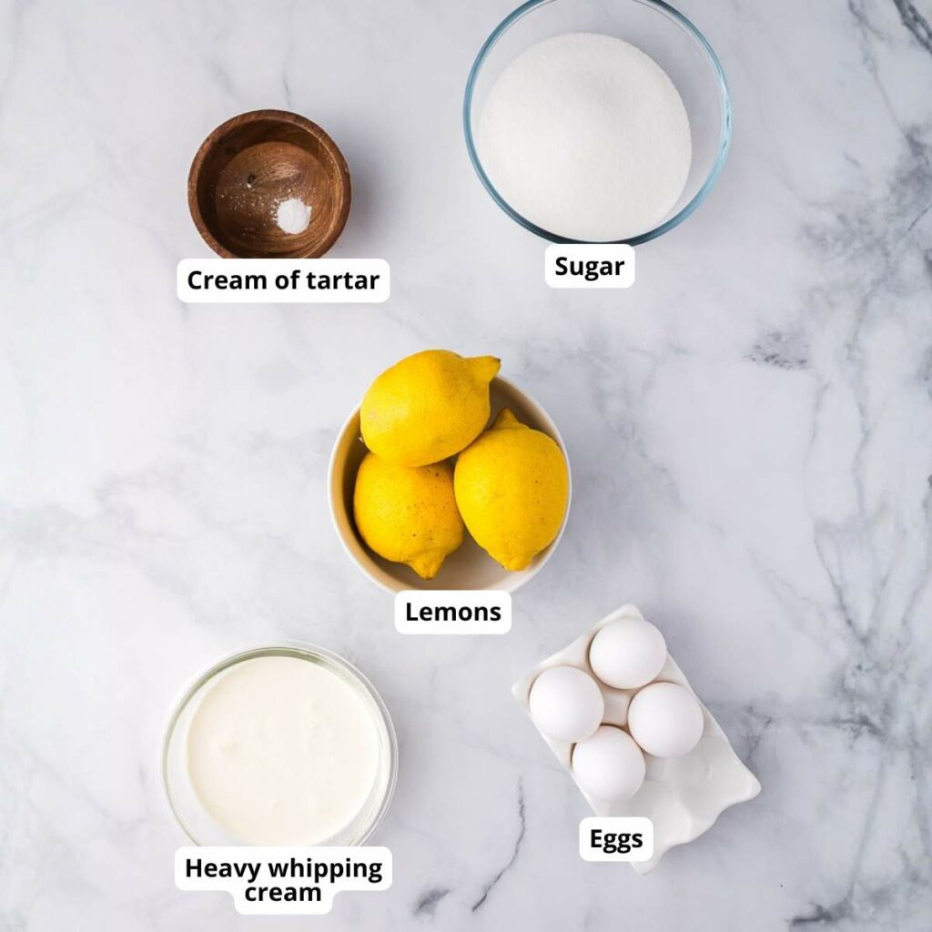 Five labeled ingredients on a marble surface: cream of tartar, sugar, lemons, heavy whipping cream, and eggs.