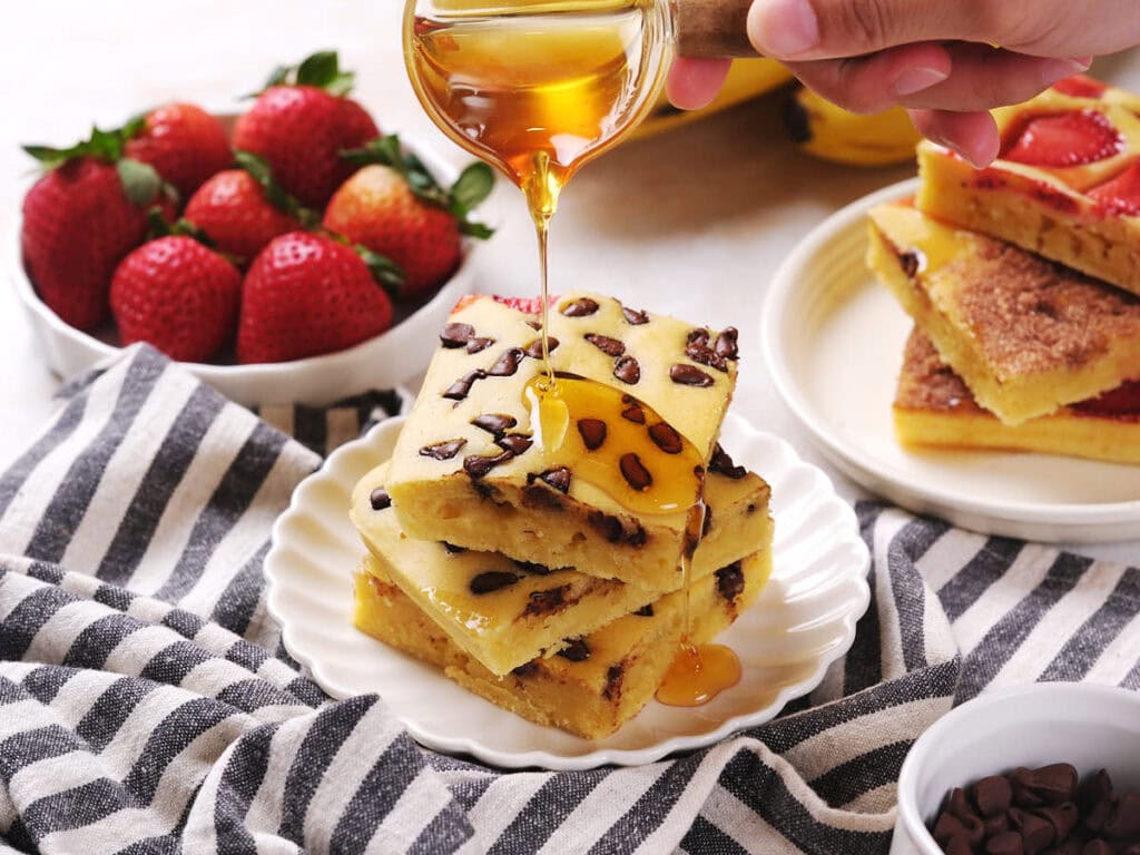 A hand pours honey over a stack of chocolate chip pancakes on a white plate, with strawberries and more pancakes in the background.