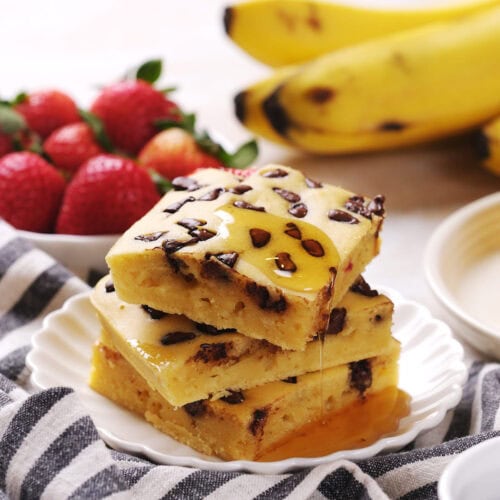 Three chocolate chip banana bars are stacked on a plate with syrup drizzled on top.