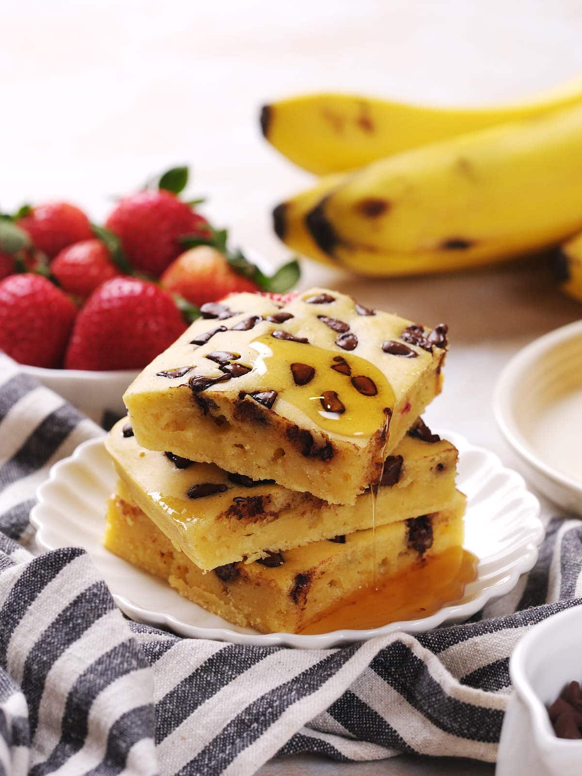 Three chocolate chip banana bars are stacked on a plate with syrup drizzled on top.