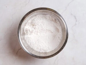 A stainless steel bowl filled with white flour sits on a light-colored marble countertop.