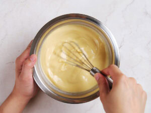 A person holds a metal bowl and whisks a creamy yellow batter or mixture on a light-colored countertop.