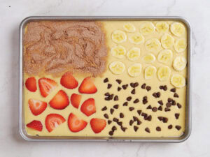 A baking sheet with batter divided into four sections, each topped with cinnamon sugar, banana slices, chocolate chips, and sliced strawberries.