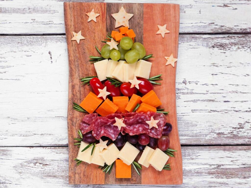 A wooden board displays cheese, grapes, tomatoes, and salami arranged in the shape of a Christmas tree.