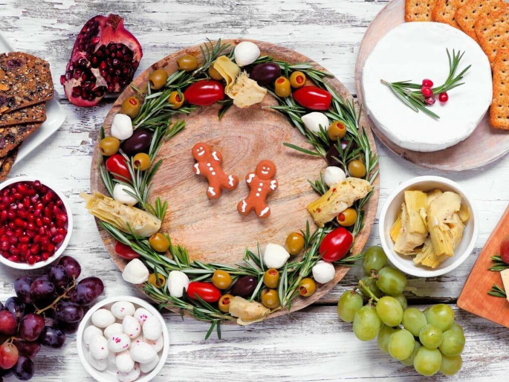 A wooden platter with a festive wreath made of tomatoes, olives, mozzarella, artichoke, and rosemary.