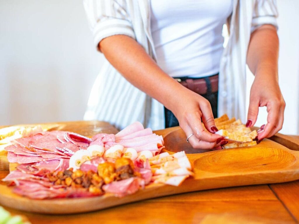 Person arranging sliced meats, cheeses, and crackers on a wooden charcuterie board.