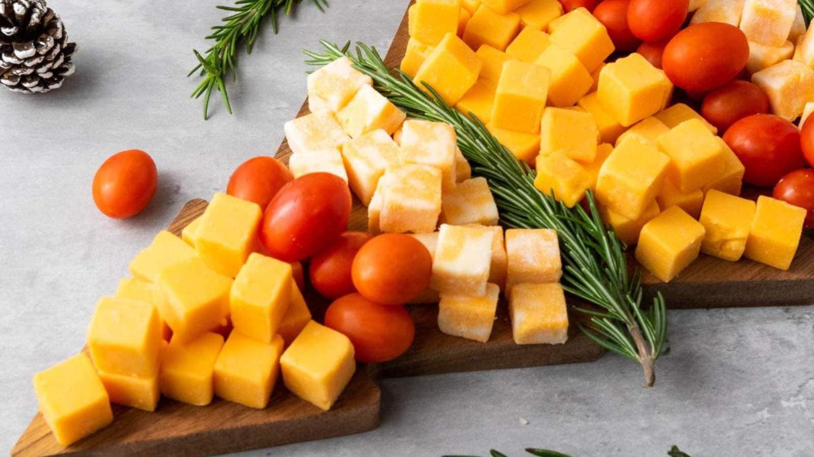 Wooden board shaped like a Christmas tree topped with cubes of yellow and white cheese, cherry tomatoes, and sprigs of rosemary.
