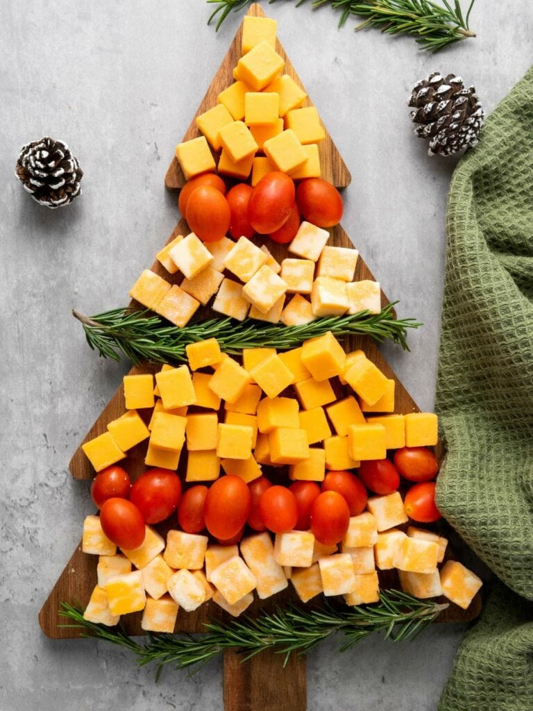 A wooden board shaped like a Christmas tree displays cubes of cheddar and colby jack cheese, grape tomatoes, and rosemary sprigs.