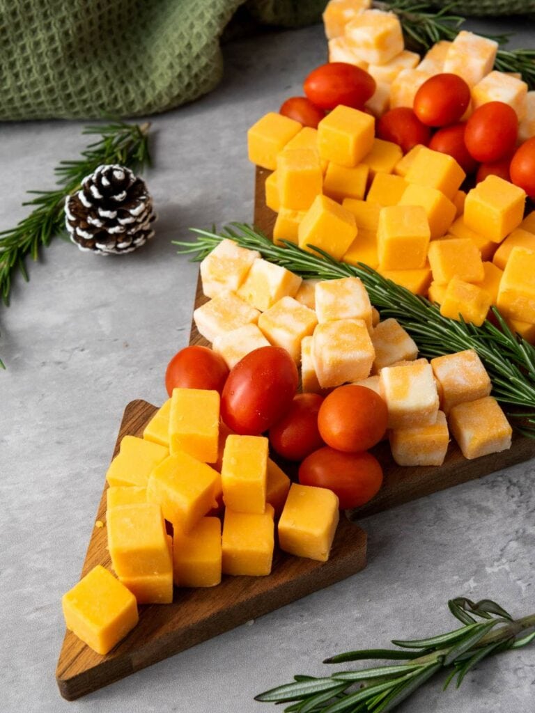 A wooden Christmas tree-shaped board with cubes of yellow and white cheese, grape tomatoes, and sprigs of rosemary.