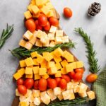 A wooden board arranged with cheese cubes, grape tomatoes, and rosemary sprigs in the shape of a Christmas tree.