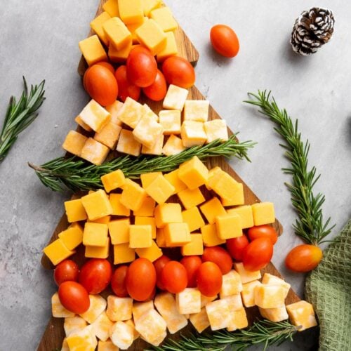 A wooden board arranged with cheese cubes, grape tomatoes, and rosemary sprigs in the shape of a Christmas tree.