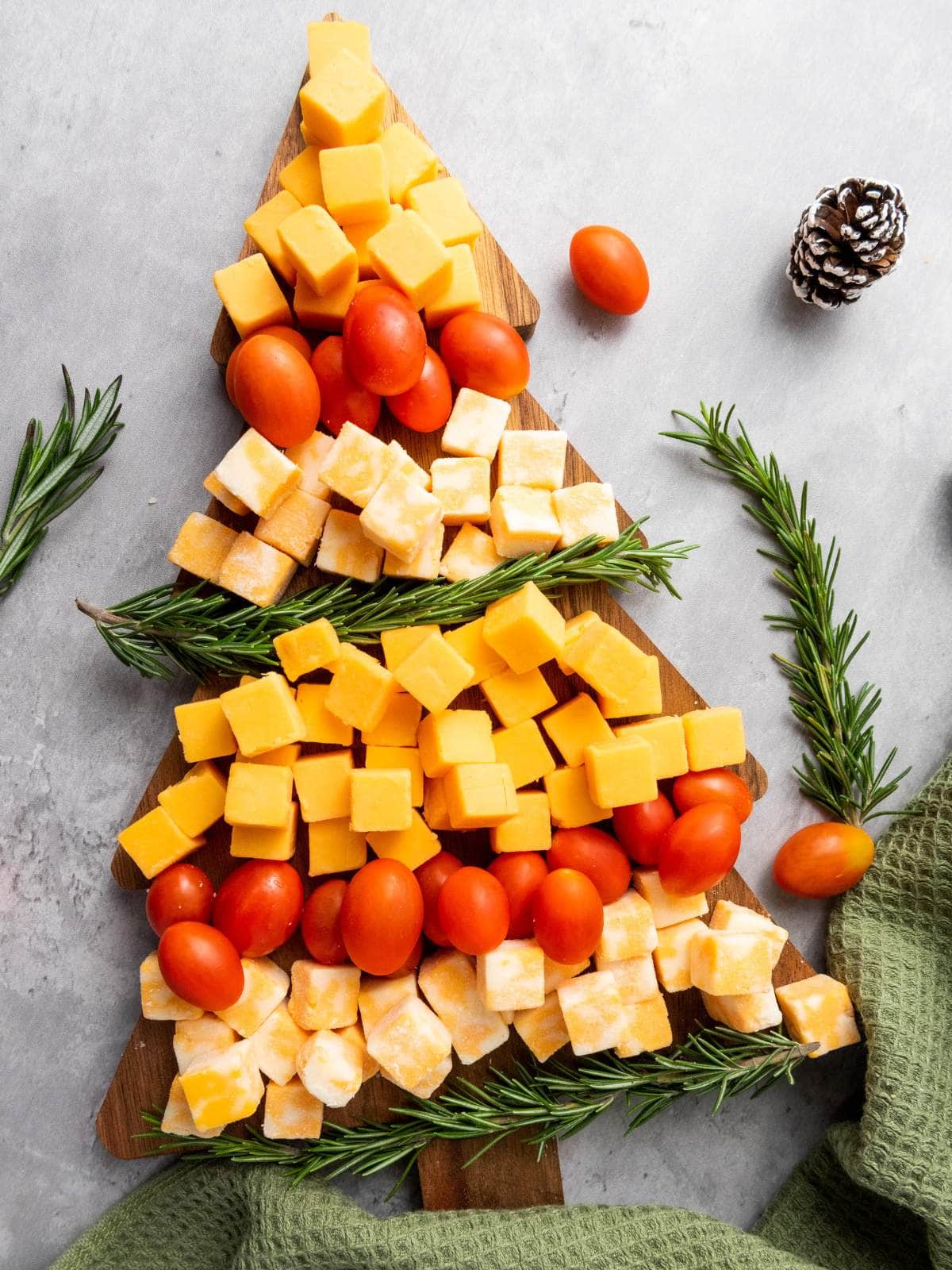 A wooden board arranged with cheese cubes, grape tomatoes, and rosemary sprigs in the shape of a Christmas tree.