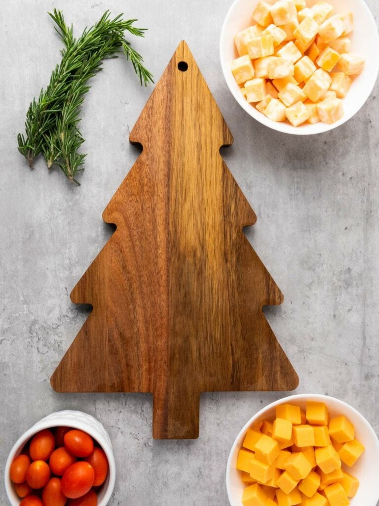 A wooden cutting board shaped like a Christmas tree is surrounded by bowls of diced cheese, grape tomatoes, and sprigs of rosemary.