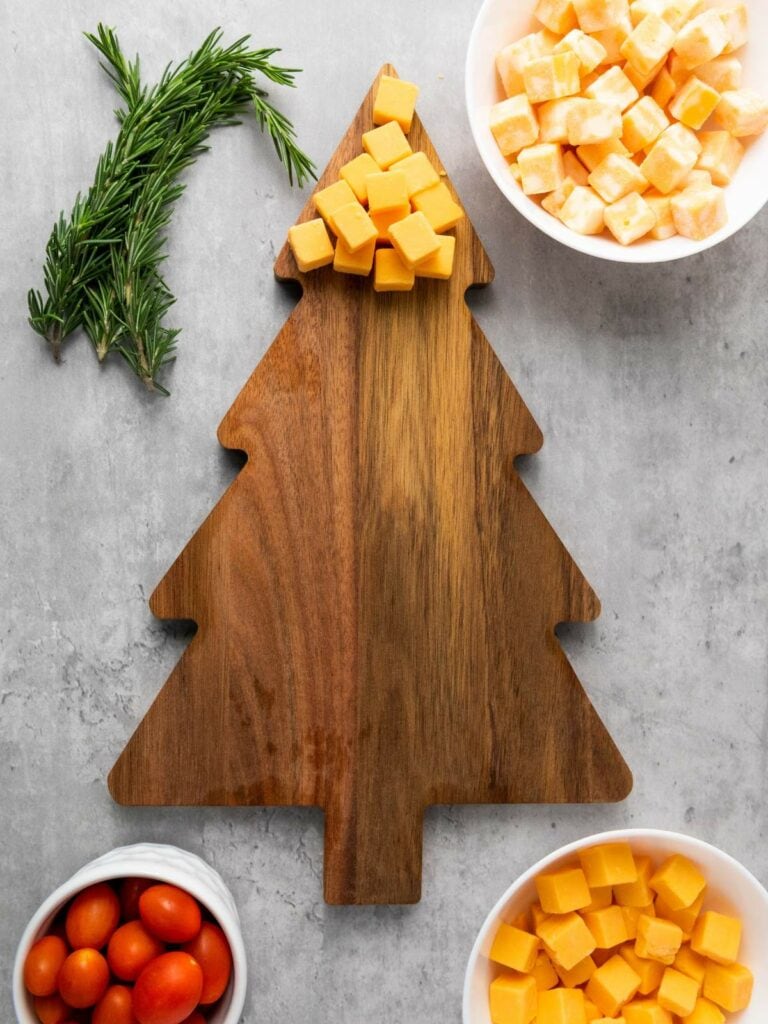 A wooden Christmas tree-shaped cutting board with cubes of cheese, grape tomatoes, and rosemary arranged nearby on a gray surface.