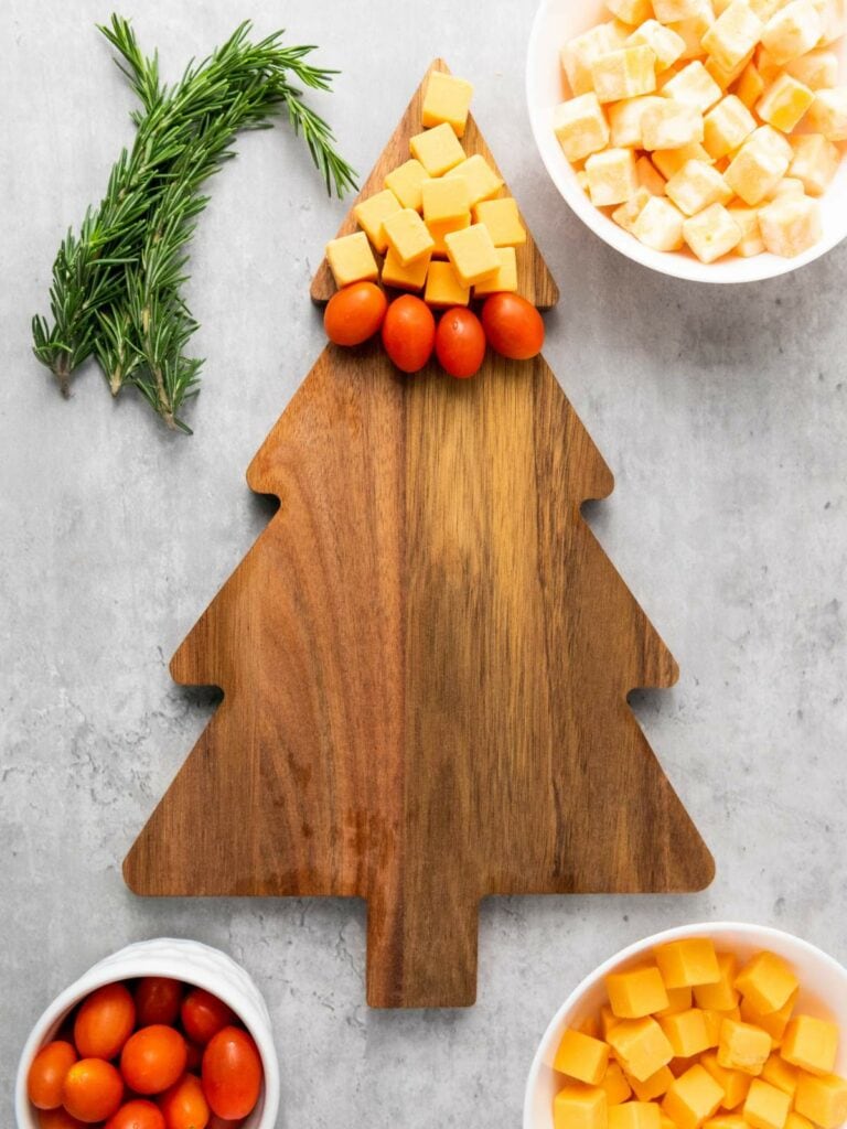 A wooden cutting board shaped like a Christmas tree with cheese cubes and grape tomatoes at the top.
