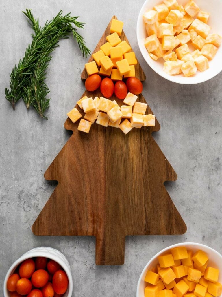 A tree-shaped wooden board with cubes of cheddar and colby jack cheese, grape tomatoes, and sprigs of rosemary arranged on a gray surface.