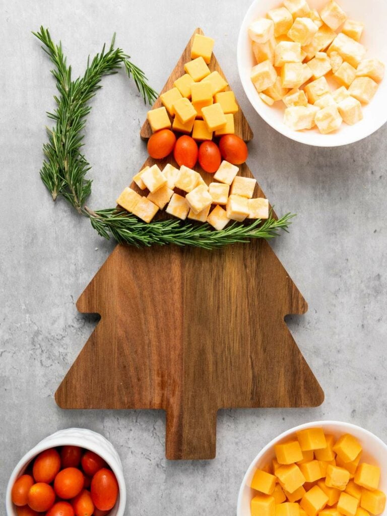 A wooden tree-shaped board with cubes of cheese, grape tomatoes, and sprigs of rosemary arranged to resemble a Christmas tree.