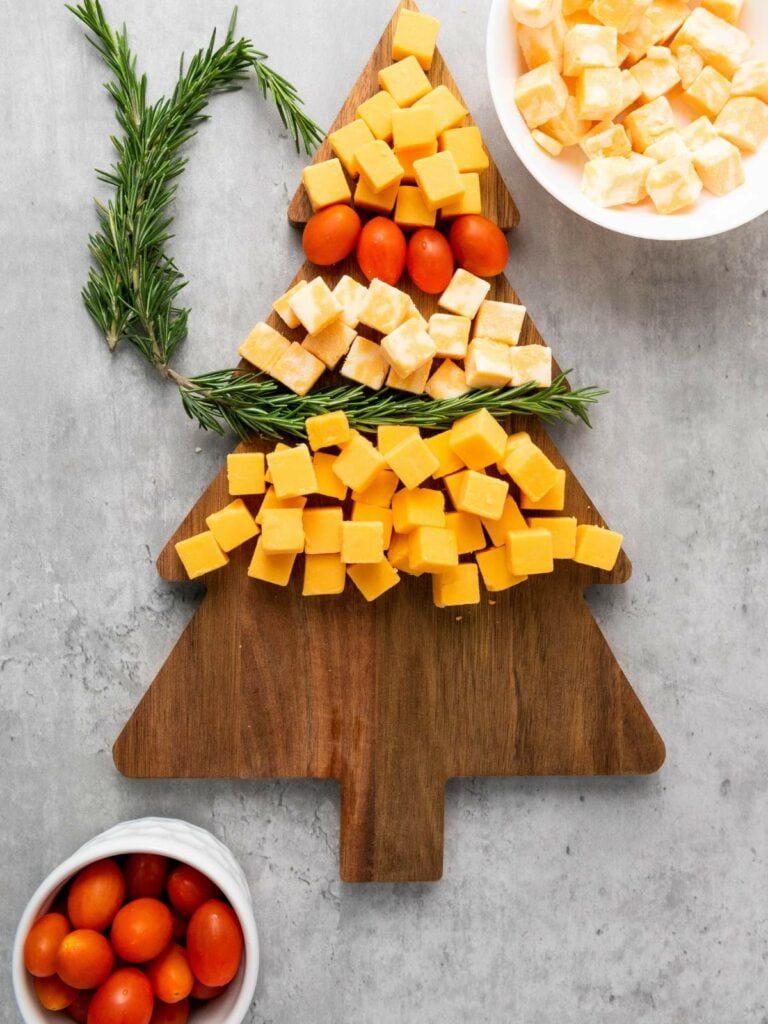 A wooden tree-shaped board with cubes of cheese, grape tomatoes, and sprigs of rosemary, with extra cheese and tomatoes in bowls nearby.