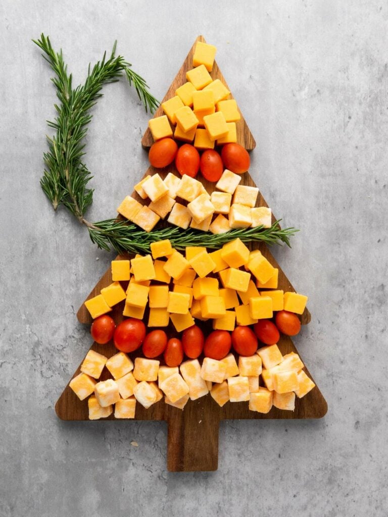 A wooden board shaped like a Christmas tree, topped with cubed cheese, cherry tomatoes, and sprigs of rosemary for decoration.