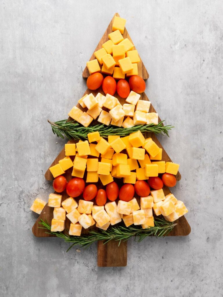 Christmas tree-shaped board with layers of cubed cheese, cherry tomatoes, and sprigs of rosemary.