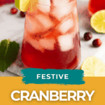 A glass of cranberry lime spritzer with ice, garnished with lime, cranberries, and mint, sits on a table with lime slices and mint leaves nearby.