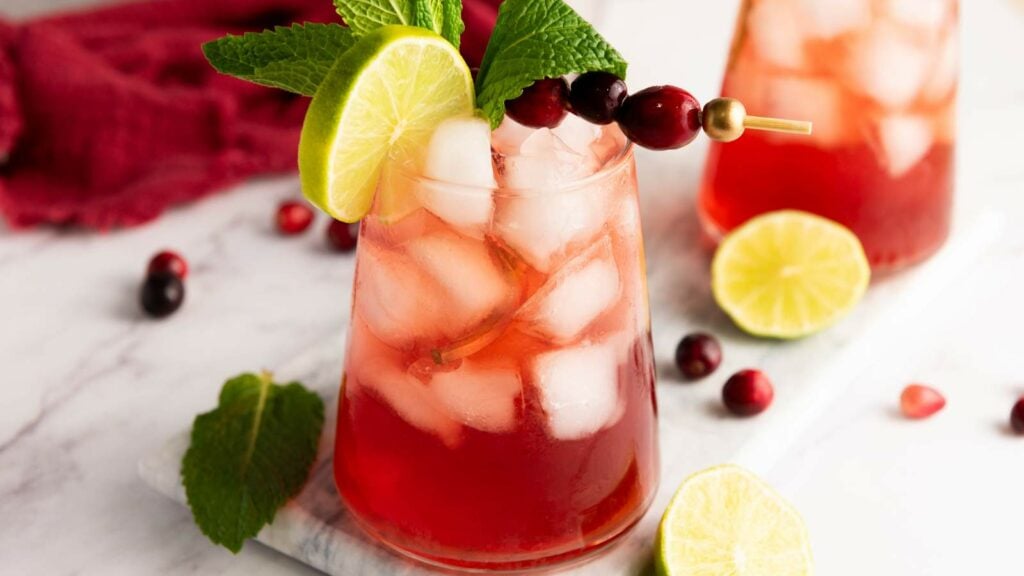 A glass of cranberry lime spritzer garnished with a lime slice, mint leaves, and cranberries.