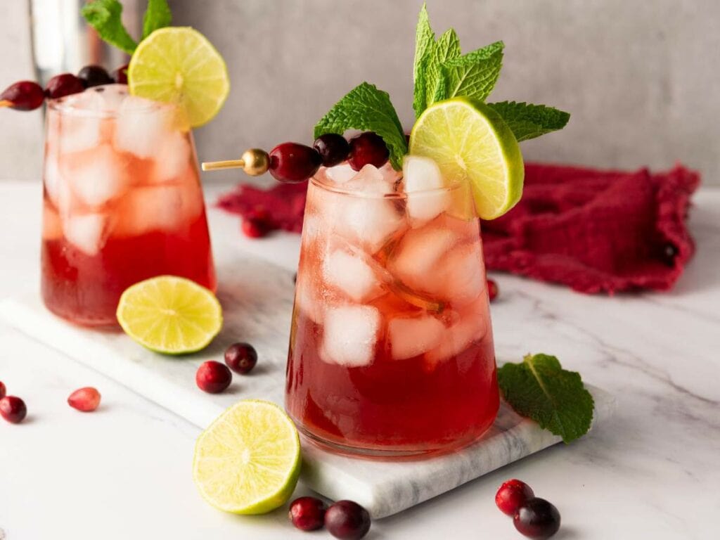Two glasses of iced cranberry cocktail garnished with lime slices, cranberries, and mint leaves on a white marble surface.