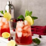 A glass of red cocktail with ice, garnished with a lime slice, cranberries, and mint, sits on a marble surface.
