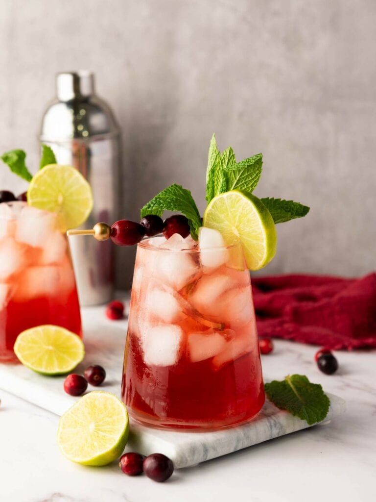 A glass of red cocktail with ice, garnished with a lime slice, cranberries, and mint, sits on a marble surface.