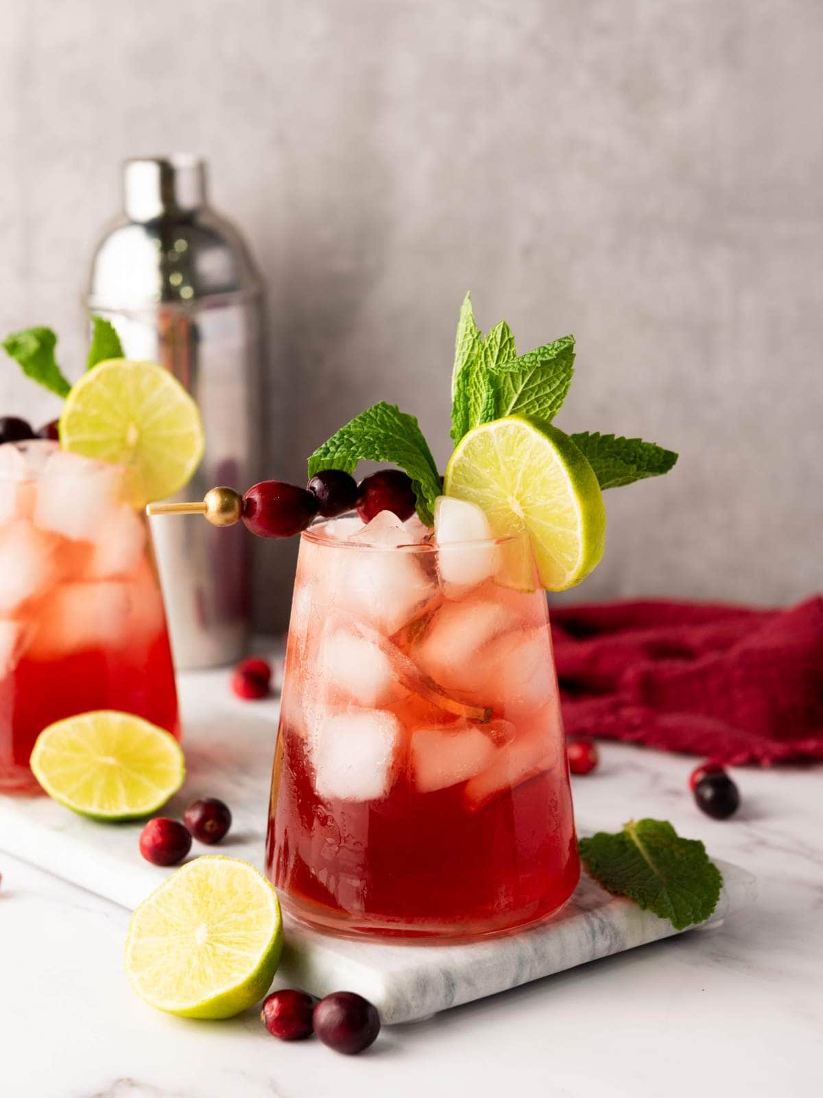 A glass of red cocktail with ice, garnished with a lime slice, cranberries, and mint, sits on a marble surface.