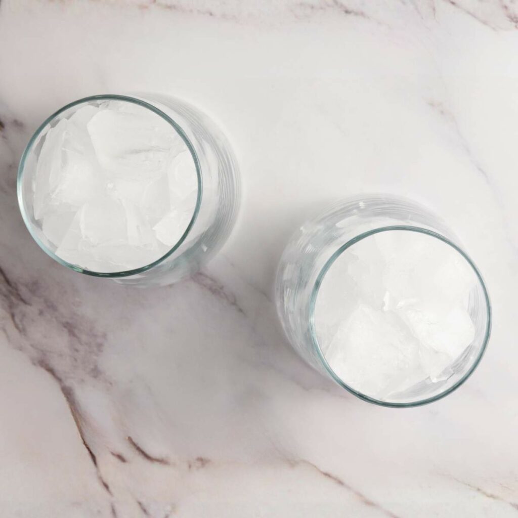 Two glass cups filled with ice cubes are placed on a white marble surface, viewed from above.