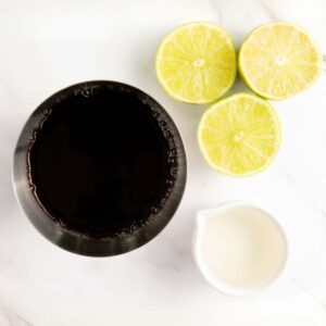 A metal cup filled with a dark liquid, a small white pitcher, and three lime halves arranged on a white surface.