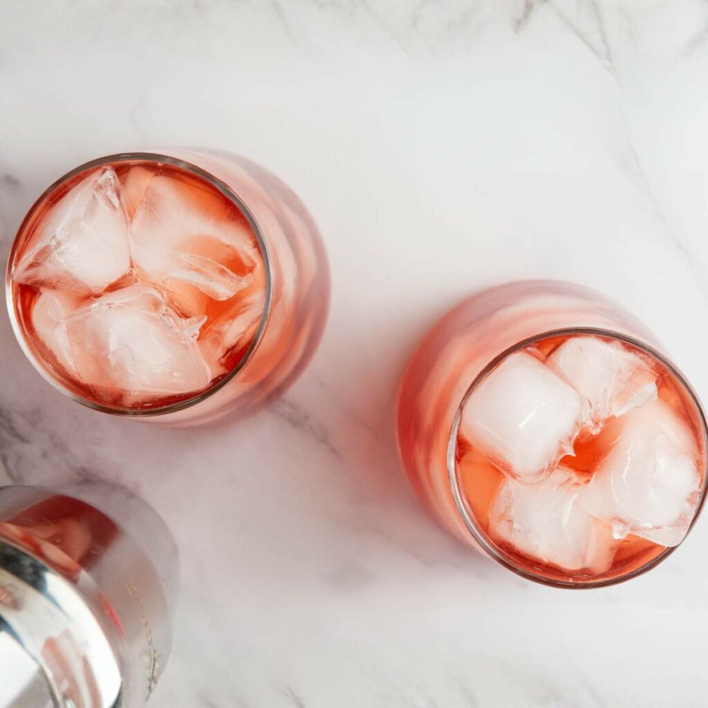 Two glasses filled with pink iced drinks sit on a white marble surface, with part of a metal cocktail shaker visible in the corner.