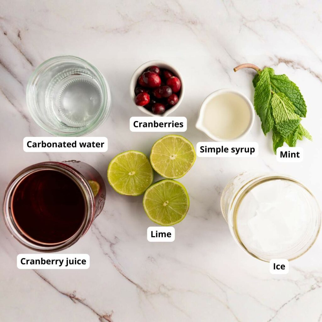 Ingredients on a white surface: carbonated water, cranberries, simple syrup, mint, lime halves, cranberry juice, and ice.