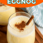 A glass of eggnog topped with ground cinnamon and a cinnamon stick, served on a wooden tray with another glass in the background.