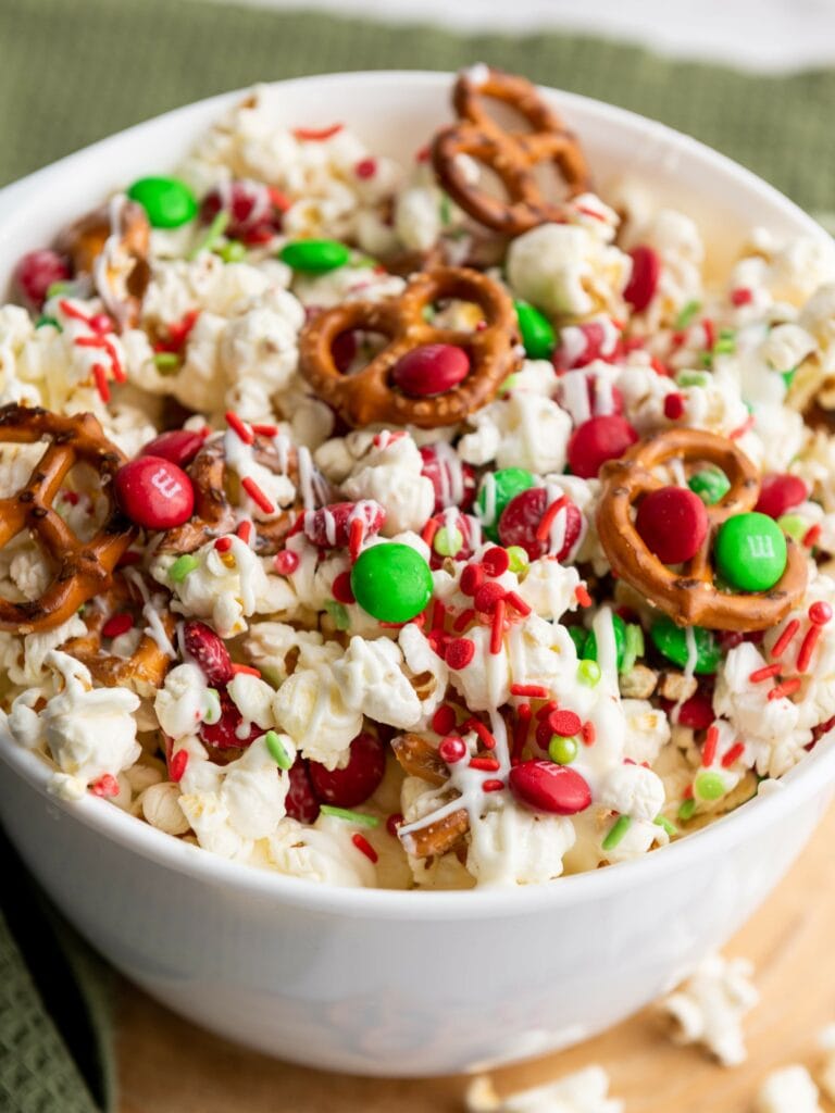 A bowl filled with popcorn, pretzels, red and green candies, red sprinkles, and drizzled white chocolate, sitting on a green cloth.