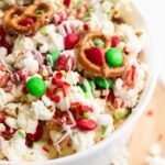 A bowl of popcorn mixed with red and green candies, mini pretzels, and red and green sprinkles, drizzled with white chocolate.
