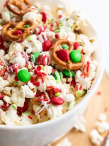 A bowl of popcorn mixed with red and green candies, mini pretzels, and red and green sprinkles, drizzled with white chocolate.