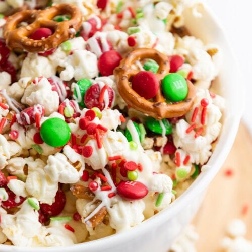 A bowl of popcorn mixed with red and green candies, mini pretzels, and red and green sprinkles, drizzled with white chocolate.