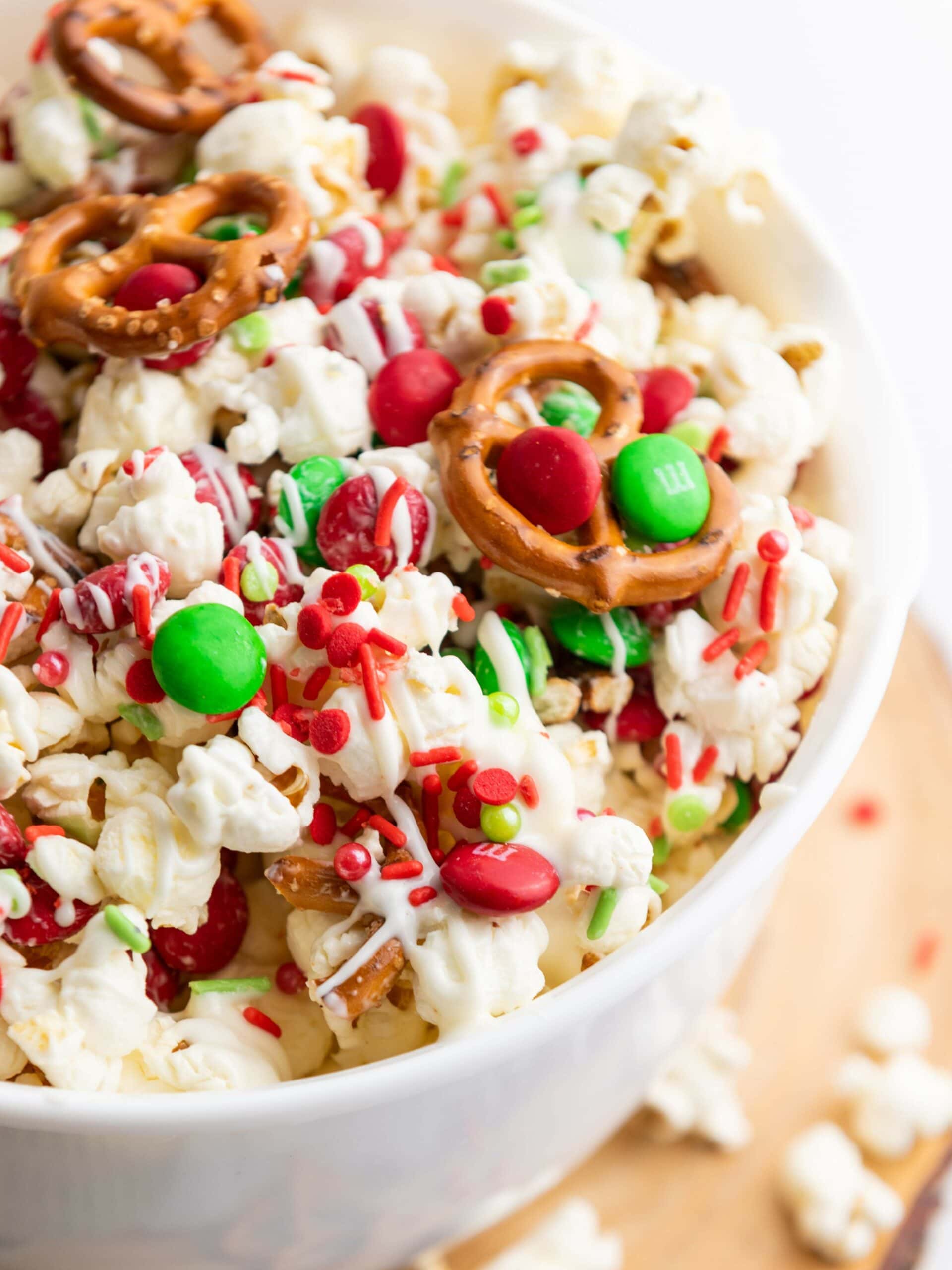 A bowl of popcorn mixed with red and green candies, mini pretzels, and red and green sprinkles, drizzled with white chocolate.