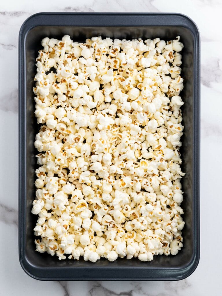 A rectangular baking pan filled with freshly popped popcorn, placed on a marble surface.