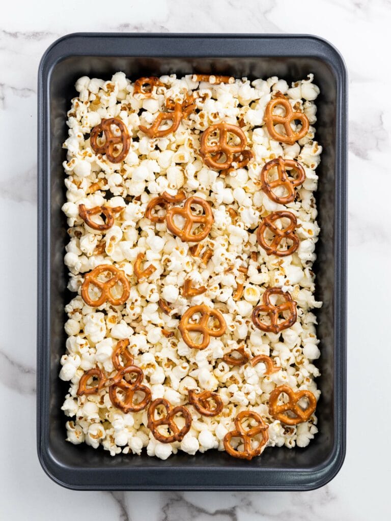 A rectangular baking pan filled with popcorn and scattered mini pretzels on a white marble surface.