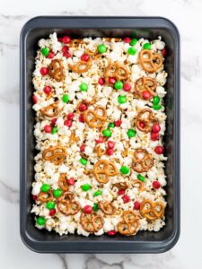 A baking pan filled with popcorn, pretzels, and red and green candy-coated chocolates on a marble surface.