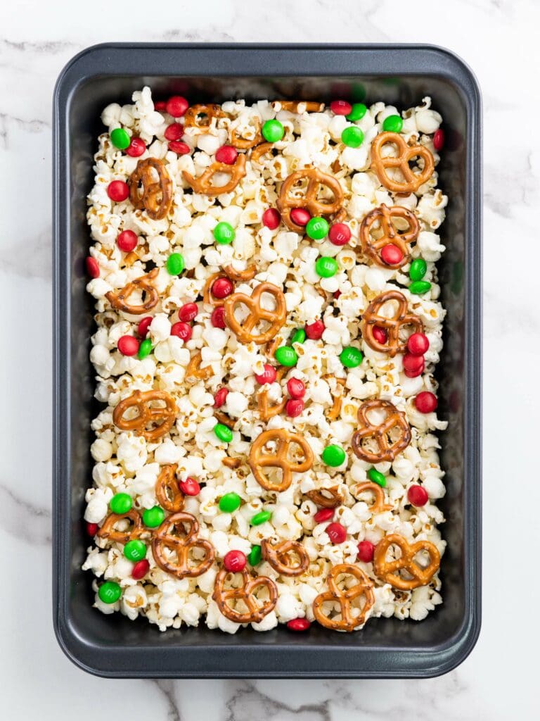 A baking pan filled with popcorn, pretzels, and red and green candy-coated chocolates on a marble surface.